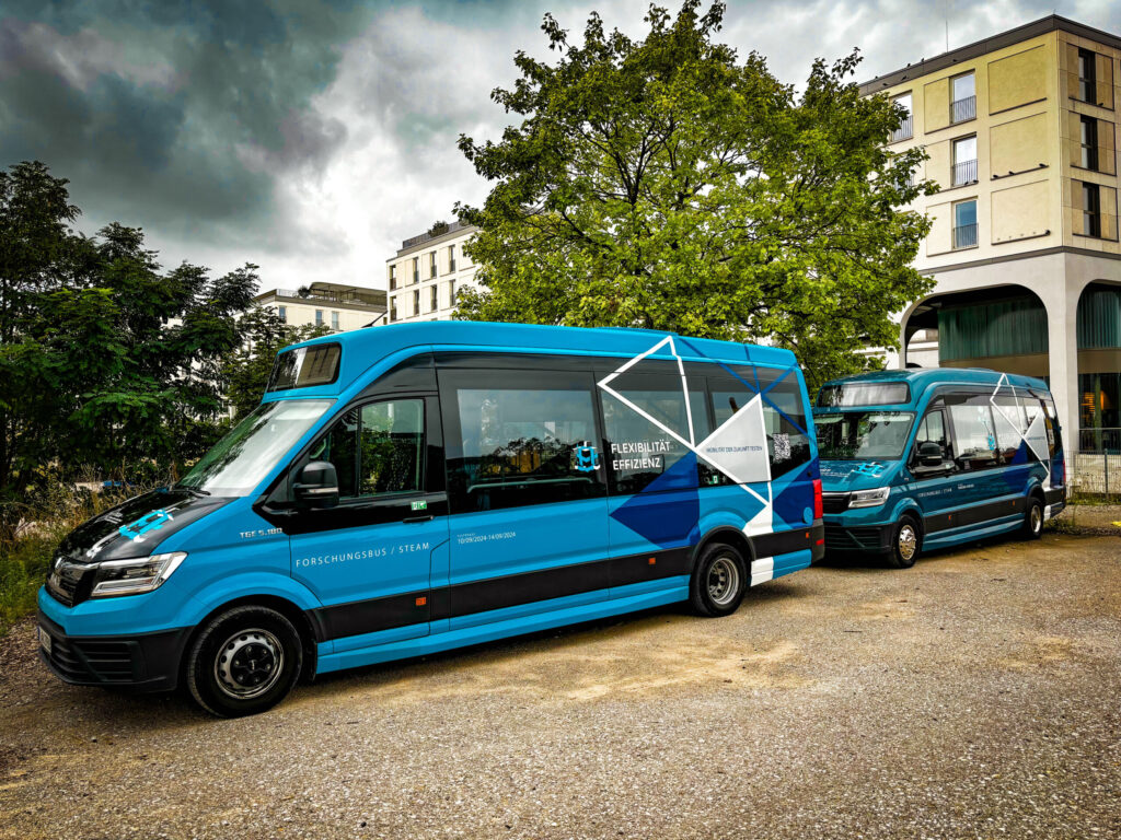 Zwei blaue Forschungsbusse mit dem Schriftzug „Forschungsbus / STEAM“ und dem Motto „Flexibilität + Effizienz“ stehen auf einem Parkplatz vor modernen Wohngebäuden und Bäumen.